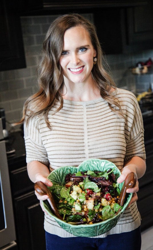 Katie Lovitt smiles at the camera as she stands in the kitchen and holds up a large bowl filled with a delicious, healthful salad | Gut Health Nutrition