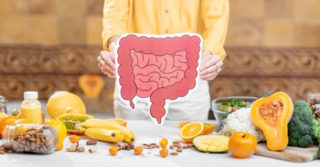 woman in yellow shirt holding a cut out image of the small and large intestine with a table in front of her filled with yellow and green fruits and vegetables | Digestive Enzymes and IBS