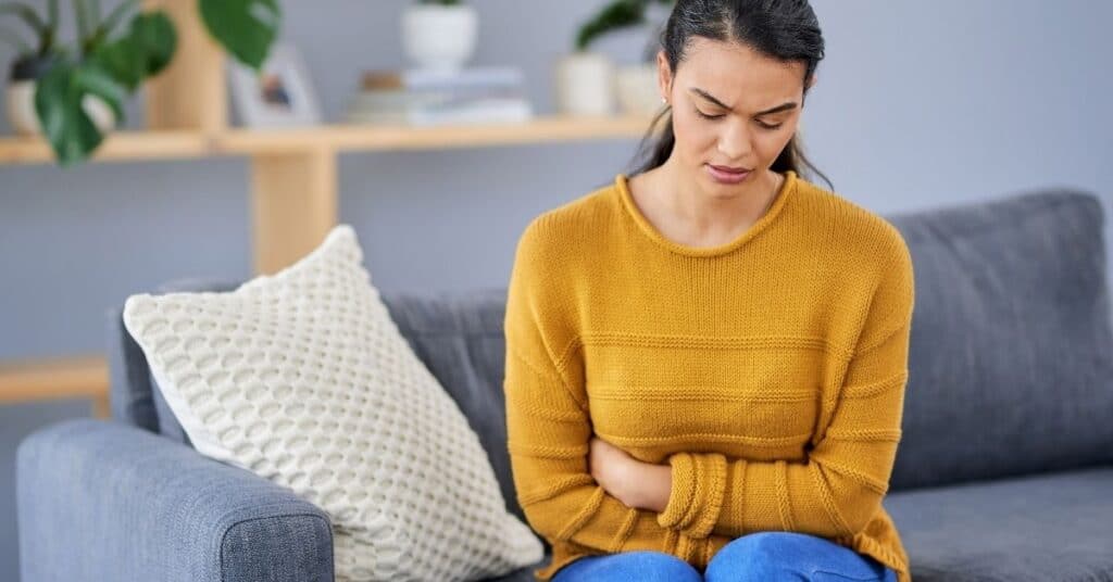 A woman sitting on a couch holding her stomach due to presumed digestive issues with a stressed look on her face