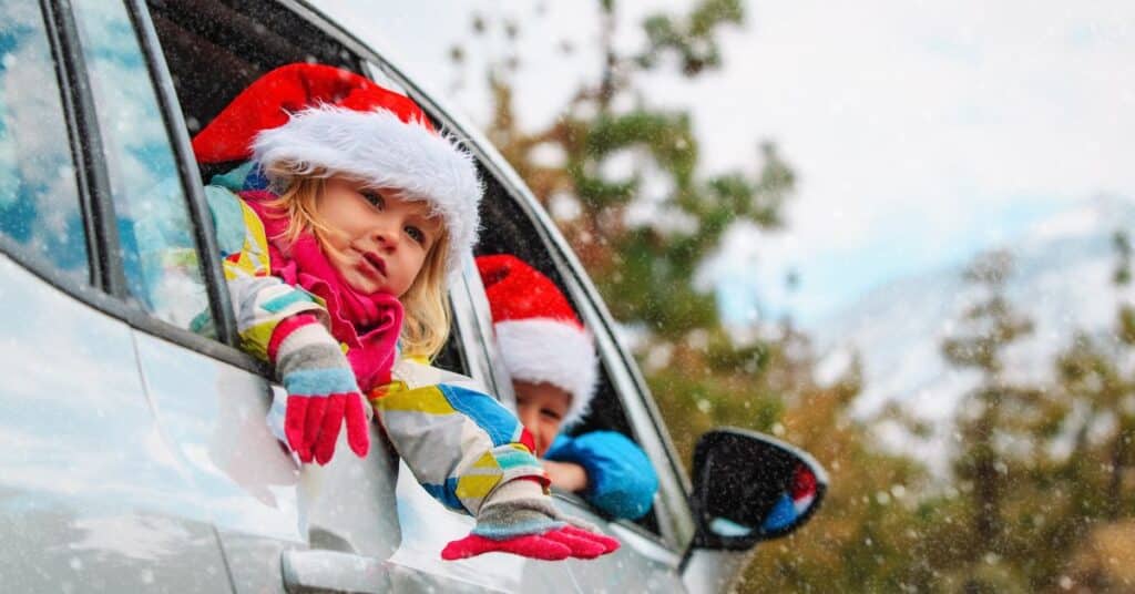 Two kids in Santa hats and gloves peer out of a car window at the snowflakes falling outside