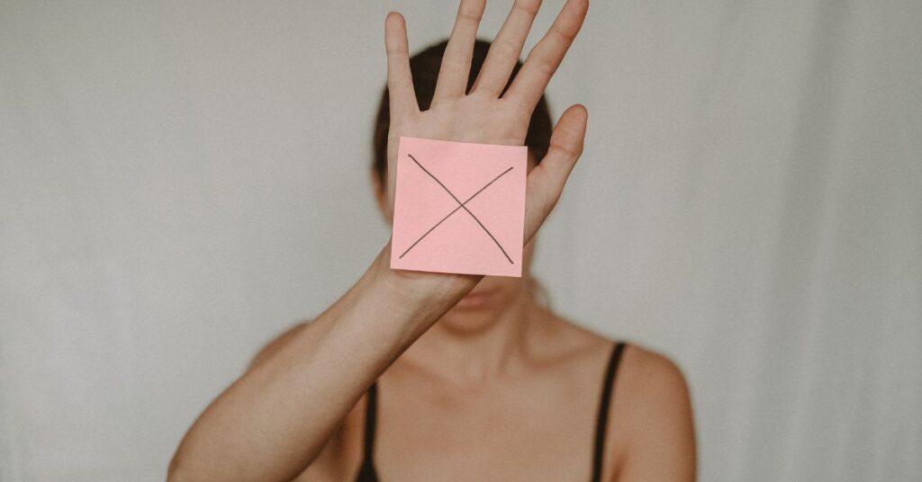 A woman holds up a sticky note with a big X on it blocking her face