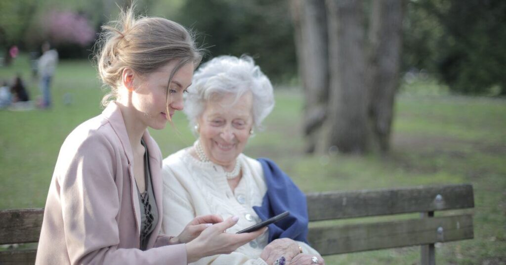 A young woman sits with an old white haired woman on a bench and shows her something on her phone