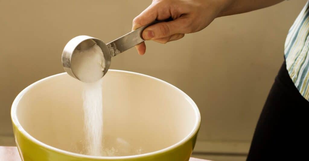 A measuring cup of sugar is dumped into a large mixing bowl