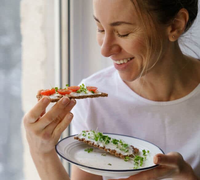 A healthy and happy woman eats veggies and soft cheese on a seed cracker, gut health virtual nutrition