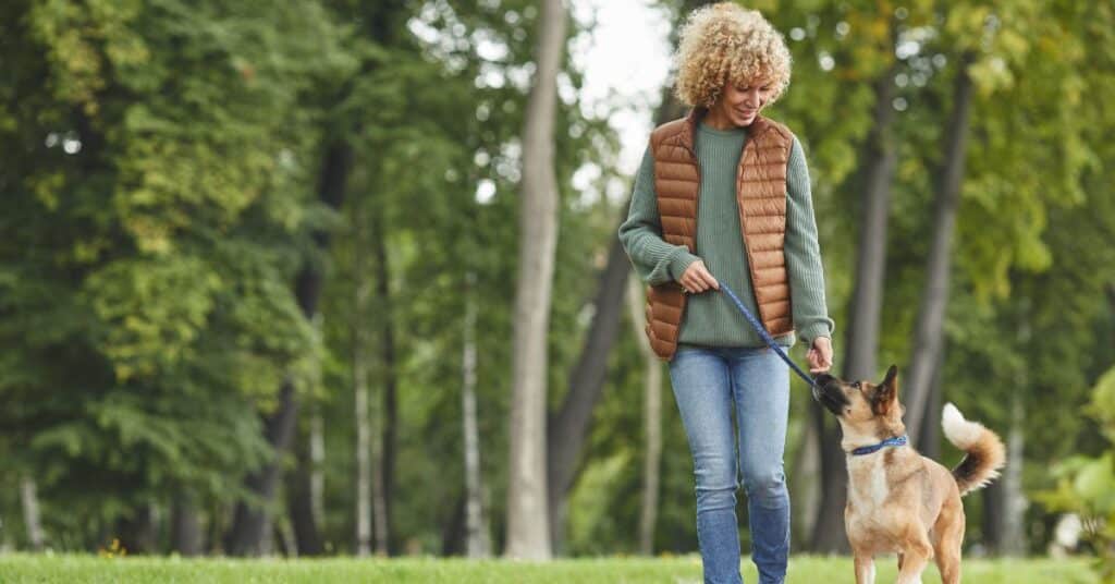 A woman in a sweater and puffy vest walks her dog outside in a natural area