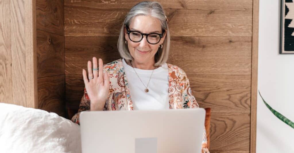 A grey-haired woman in glasses waves high to her computer on a video call, hiring help for reducing IBS symptoms