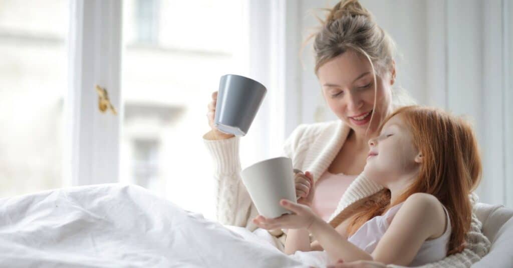 A mom and daughter snuggle under some blankets with mugs of a warm beverage