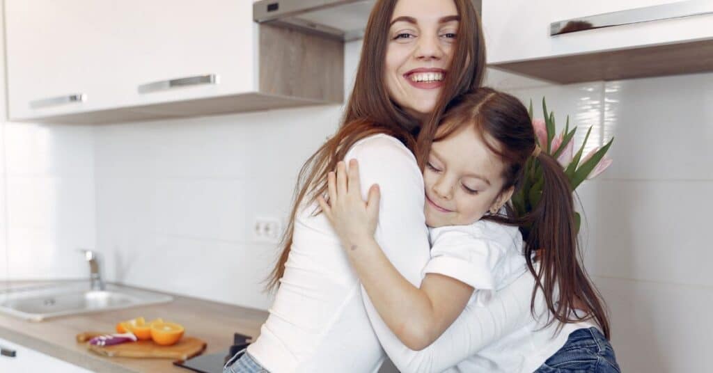 A mom smiles wide as she hugs her young daughter in a modern kitchen, digestive symptoms