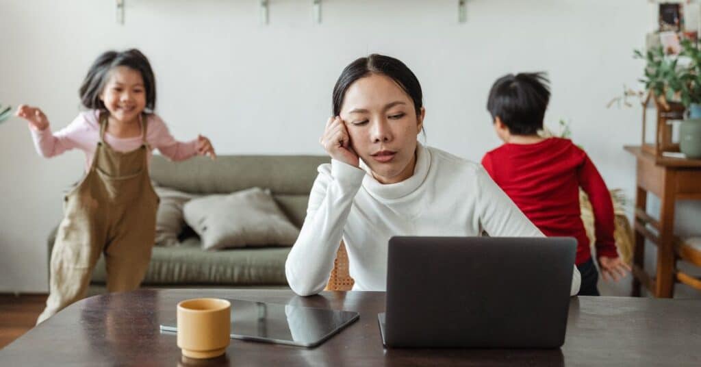 A mom tries to ignore her playing children in the background as she works on a laptop, hot mess mom culture