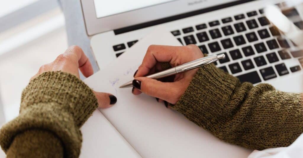 A woman writes in a notebook while working at a laptop